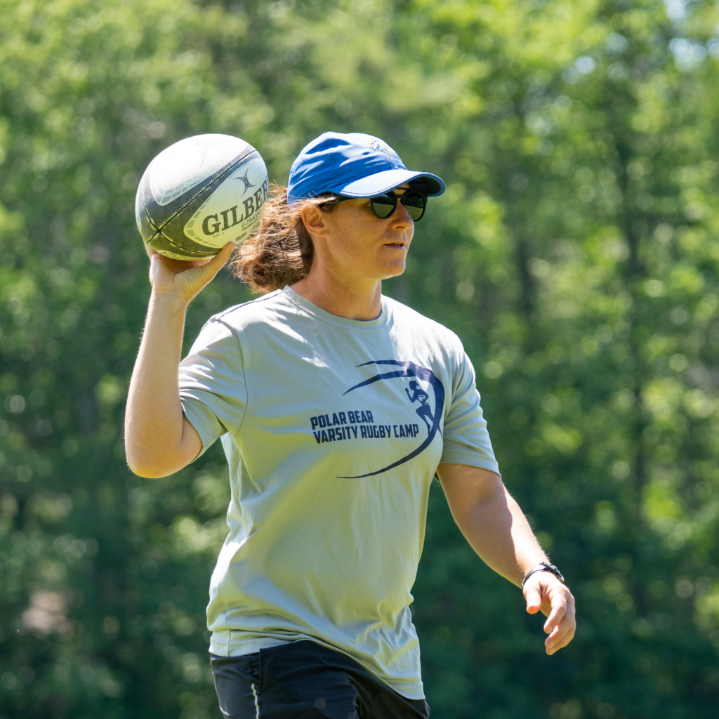 A coach in a baseball cap holding up a rugby ball
