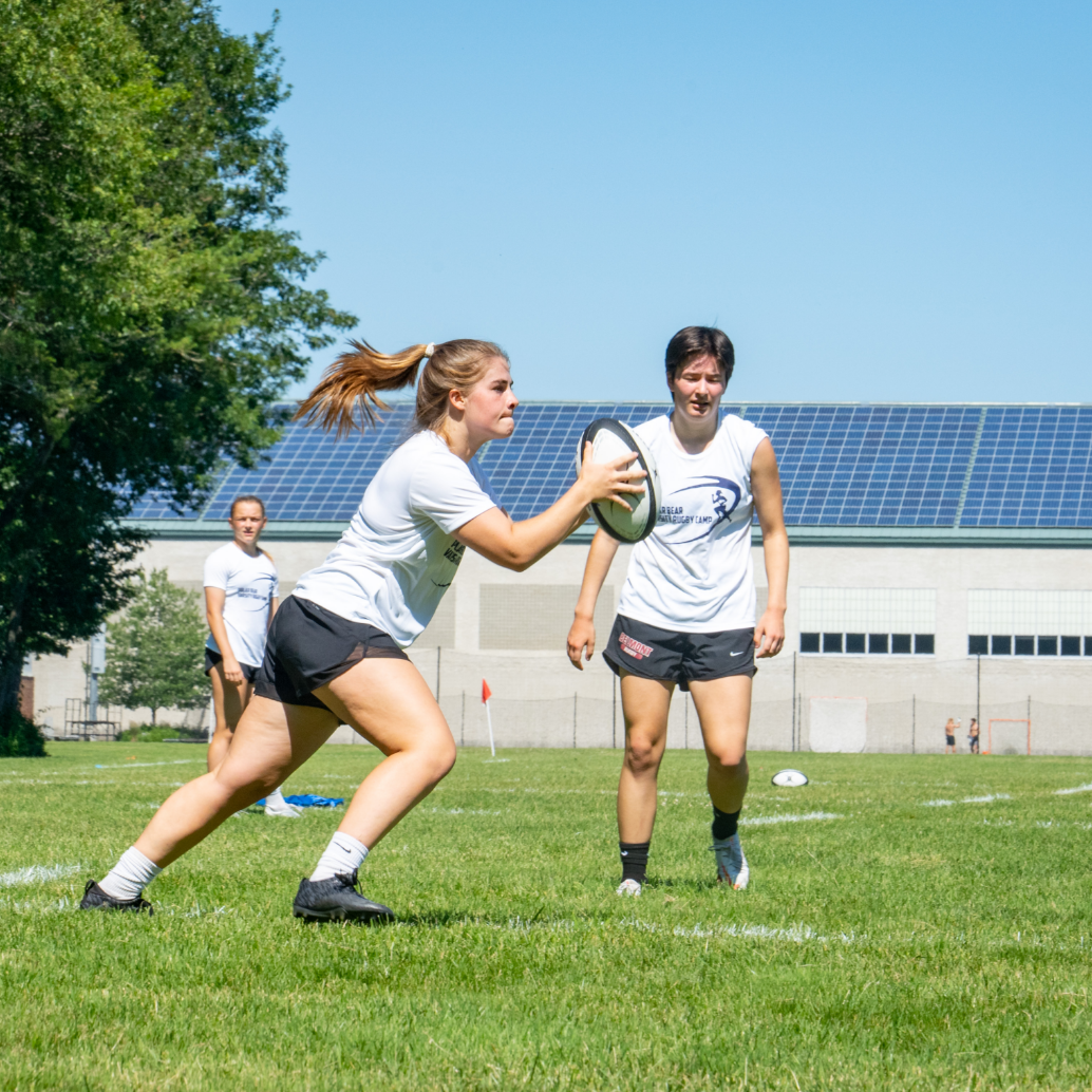 One girl running with a rugby ball while another in the background looks on